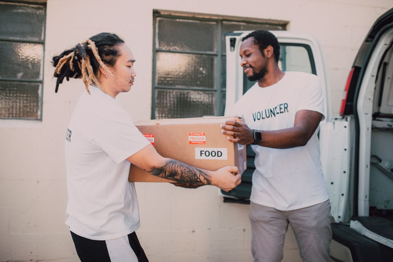 Free Two volunteers transferring a food box from a van, symbolizing community support and charity work. Stock Photo